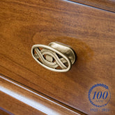 Dettaglio di un pomello in ottone decorato su un mobile in legno lucido color ciliegio, con incisioni eleganti in stile classico. Nell’angolo in basso a destra, un logo celebra i 100 anni dell’azienda (1925–2025) con una corona d’alloro e la scritta “100 anni